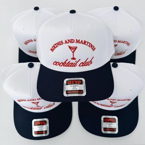 May include: Five white and blue baseball caps with the text "Bikinis and Martinis cocktail club" and a martini glass graphic in red. The caps have a navy blue brim and a white crown.