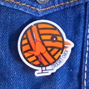 May include: A round, white pin featuring an orange and yellow yarn ball with a crochet hook and a bird-like face. The text "CROCHET CHICK" is printed on the pin. The pin is set against a blue denim background.