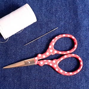 May include: A spool of white thread, a needle, and a pair of red scissors with white polka dots rest on a blue denim surface. The scissors are open, ready for use. This image is related to sewing and crafting.