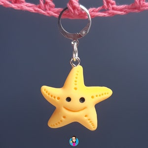 Pode incluir: Um amuleto de estrela do mar amarela sorridente com um chaveiro prateado. O amuleto está pendurado em um cordão de crochê rosa.