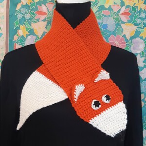 May include: A crocheted orange and white fox scarf. The scarf is designed to look like a fox, with a white tail and a fox head with black and white eyes. The scarf is displayed on a black shirt.