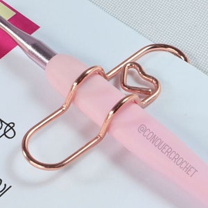 May include: A pink crochet hook with a gold heart-shaped hook holder. The hook is labelled "5/0 3.0mm" and "@CONQUER.CROCHET".