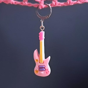 K&ouml;nnte beinhalten: Ein rosa Gitarren-Anh&auml;nger mit einem gelb-wei&szlig; gestreiften Hals, violetten und goldenen Akzenten und einem silbernen Haken.