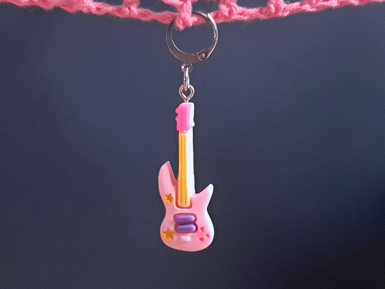 K&ouml;nnte beinhalten: Ein rosa Gitarren-Anh&auml;nger mit einem gelb-wei&szlig; gestreiften Hals, violetten und goldenen Akzenten und einem silbernen Haken.