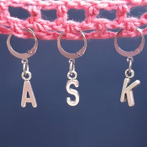 May include: Three silver letter charms with the letters A, S, and K hanging from silver hoops. The charms are hanging from a pink crocheted fabric.