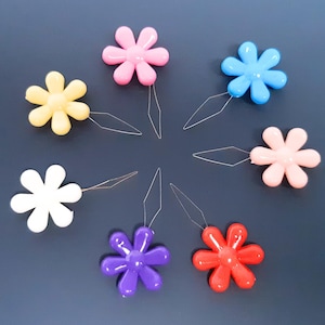 May include: A set of seven colourful flower-shaped threaders with a needle attached to each. The threaders are pink, yellow, white, blue, peach, purple, and red.