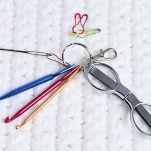 May include: A set of crochet hooks, a pair of small scissors, and a keyring with a colourful paperclip attached. The items are on a white crocheted surface.