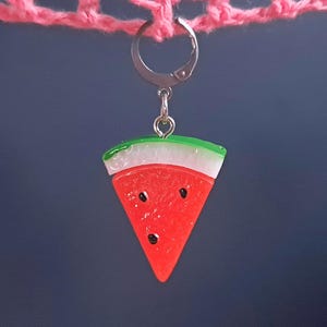 May include: A watermelon slice charm with a green rind, white flesh, and red interior with black seeds. The charm is attached to a silver keyring.