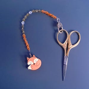May include: Gold embroidery scissors with a fox charm. The charm features a sleeping fox in orange and white, connected to a beaded chain. The scissors have ornate handles and silver blades. The background is a dark blue.