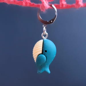 May include: A blue and cream-colored whale charm hangs from a silver hoop. The whale has a rounded body with a textured cream-colored head and a blue body. The charm is suspended against a dark blue background.