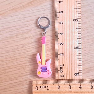 K&ouml;nnte beinhalten: Rosa-gelber Gitarren-Schl&uuml;sselanh&auml;nger mit einem silbernen Schl&uuml;sselring. Die Gitarre hat ein lila-gelbes Design mit Sternen und einem rosa-wei&szlig;en Schlagbrett. Der Schl&uuml;sselanh&auml;nger ist etwa 6 cm lang.