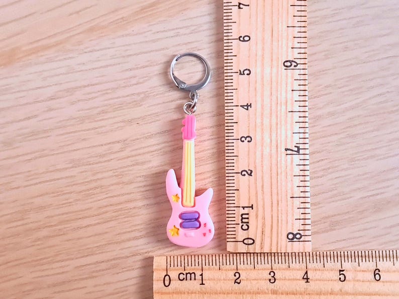 K&ouml;nnte beinhalten: Rosa-gelber Gitarren-Schl&uuml;sselanh&auml;nger mit einem silbernen Schl&uuml;sselring. Die Gitarre hat ein lila-gelbes Design mit Sternen und einem rosa-wei&szlig;en Schlagbrett. Der Schl&uuml;sselanh&auml;nger ist etwa 6 cm lang.