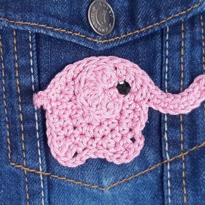 Puede incluir: Una aplicación de elefante de crochet rosa con un ojo negro, unida a una chaqueta vaquera azul.
