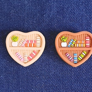May include: Two heart-shaped decorations, each depicting a bookshelf. One is peach, the other brown. Each shelf is filled with colourful books and a small potted plant, creating a miniature library theme.