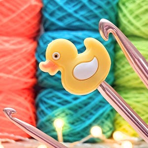 May include: A yellow rubber duck-shaped crochet hook handle attachment featuring a white wing detail and a black eye. The duck is attached to a metallic silver crochet hook, with blurred colourful yarn balls in the background.