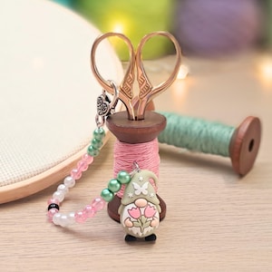 May include: Gold-coloured embroidery scissors sit on a pink thread spool, with a beaded charm of a gnome holding flowers. A second spool of green thread is in the background. The image evokes a crafting or sewing theme.