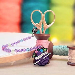 May include: Gold-toned scissors resting in a wooden spool, attached to a beaded chain with purple, blue, and green crystals. A circular enamel pin features a purple yarn ball, a white crochet hook, and the text CROCHETALLDAY with floral accents.