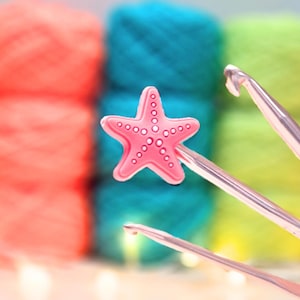 May include: A pink starfish-shaped crochet hook handle topper featuring a series of small circular indentations along each arm, attached to a metallic silver crochet hook, with blurred colourful yarn skeins in the background.