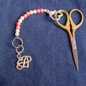 May include: A gold pair of small scissors with a pink and white beaded keychain attached. The keychain has a silver charm in the shape of a heart.
