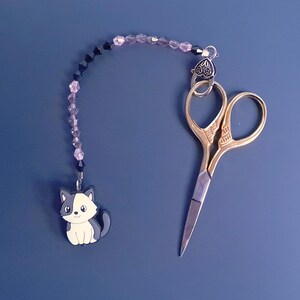 May include: A black and white cat charm hangs from a beaded chain with a pair of gold scissors attached to the other end. The chain has black and pink beads.