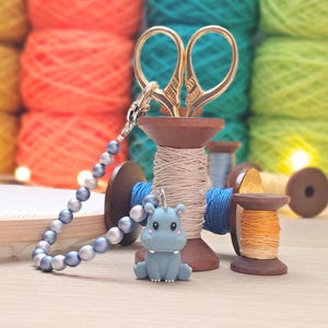 May include: A craft-themed still life featuring a blue hippo keyring, gold scissors, and spools of thread. A beaded bracelet and embroidery hoop are also present. Colourful yarn in shades of orange, green, and yellow fill the background.
