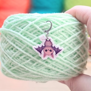May include: A light green ball of yarn with a bat-shaped stitch marker attached. The bat is purple and pink with a silver-colored metal loop. The yarn is held in a hand, with other balls of yarn in the background.