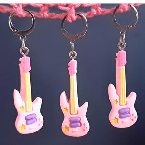 K&ouml;nnte beinhalten: Drei rosafarbene Gitarren-Ohrringe mit silbernen Haken. Jede Gitarre hat eine gelbe Saite, violette Details und rosa Sterne.
