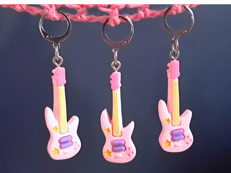 K&ouml;nnte beinhalten: Drei rosafarbene Gitarren-Ohrringe mit silbernen Haken. Jede Gitarre hat eine gelbe Saite, violette Details und rosa Sterne.