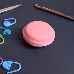 May include: A selection of crochet tools on a dark surface. The items include a pink round container, a copper crochet hook, gold scissors, and colourful stitch markers in blue, green, and purple.
