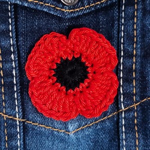Puede incluir: Una flor de amapola roja de ganchillo con un centro negro, prendida a una chaqueta vaquera azul.