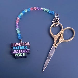 May include: A gold pair of small scissors with a beaded chain attached to a black charm with the text "I have it all together... I just can't find it".