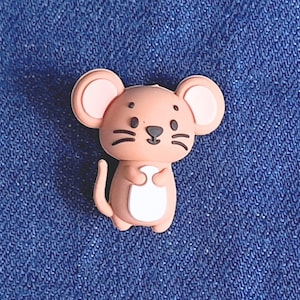 May include: A light brown mouse-shaped charm with pink ears and a white belly. The mouse has black whiskers and eyes. The charm is set against a blue denim background.
