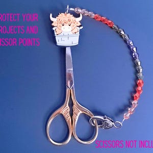 Pu&ograve; includere: Un copripunta per forbici con un'illustrazione di una mucca Highland beige in un secchio bianco, collegato a una catena di perline sfaccettate rosse e grigie. Il testo recita: PROTECT YOUR PROJECTS AND SCISSOR POINTS. SCISSORS NOT INCLUDED.