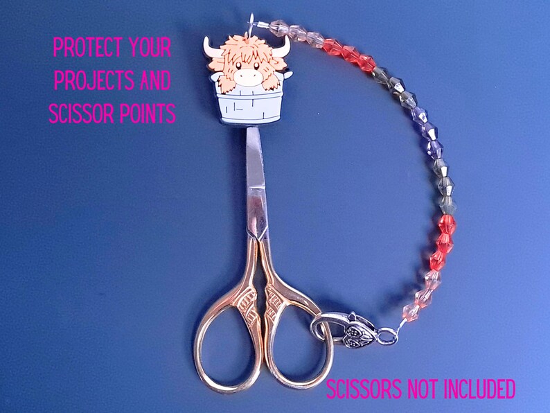 Pu&ograve; includere: Un copripunta per forbici con un'illustrazione di una mucca Highland beige in un secchio bianco, collegato a una catena di perline sfaccettate rosse e grigie. Il testo recita: PROTECT YOUR PROJECTS AND SCISSOR POINTS. SCISSORS NOT INCLUDED.