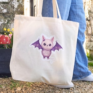 May include: A cream tote bag with a sticker of a cartoon bat with purple wings and a pink body. The bag is held by someone wearing blue jeans and white trainers. The background shows a stone wall and some greenery.