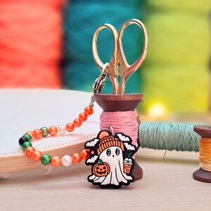May include: A ghost-shaped charm featuring a white ghost wearing an orange beanie, holding a pumpkin and a drink, attached to a beaded chain with orange, green, and white beads. The charm is displayed alongside gold scissors and wooden spools.