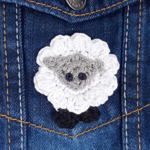 Puede incluir: Un aplique de oveja de ganchillo hecho a mano en blanco, gris y negro, fijado a una chaqueta vaquera azul. La oveja tiene un cuerpo blanco esponjoso, cabeza gris y patas negras. La chaqueta tiene un bolsillo y costuras visibles.