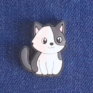 May include: A cartoon cat charm in black and white. The cat is depicted sitting with its tail curled. The charm has a black outline and is set against a blue background.