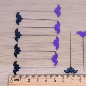 以下が含まれることがあります： 黒いコウモリの形をしたピンが9本、紫色のピンが9本、紫色のコウモリの形をしたピンが1本、黒いピンが1本。ピンは木製の表面に2列に並べられており、定規は10cmを測定しています。