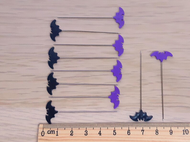以下が含まれることがあります： 黒いコウモリの形をしたピンが9本、紫色のピンが9本、紫色のコウモリの形をしたピンが1本、黒いピンが1本。ピンは木製の表面に2列に並べられており、定規は10cmを測定しています。