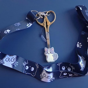 May include: A black and white cat-themed lanyard with paw prints and a gold pair of scissors attached. The lanyard has a small grey and white cat charm attached to the scissors.