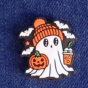 May include: A Halloween-themed charm featuring a white ghost wearing an orange beanie. The ghost is surrounded by bats, a jack-o'-lantern, and a drink. The charm is set against a blue background.