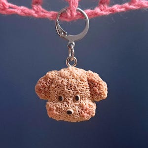 May include: A light brown dog-shaped keychain with a silver ring. The dog has a textured surface and dark eyes. The keychain is hanging from a pink crocheted string against a dark blue background.