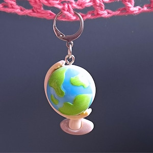 Könnte beinhalten: Ein kleiner, blauer und grüner Globus-Anhänger mit einem silbernen Ring. Der Globus steht auf einem weißen Sockel und hängt an einer rosa gehäkelten Oberfläche.