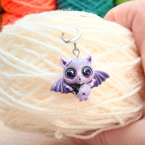 May include: A light purple bat-shaped stitch marker with large eyes and purple wings is attached to a silver-colored ring. The marker is held in front of a ball of cream-colored yarn. Colorful yarn is visible in the background.