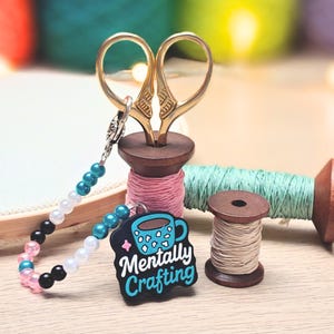 May include: A selection of craft items, featuring gold scissors, pink, green, and beige thread spools, and a beaded charm with a "Mentally Crafting" tag. The scissors are placed in a pink thread spool. The thread spools are in cm.