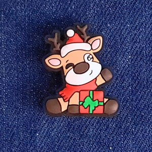 May include: A festive charm depicting a winking reindeer wearing a Santa hat and red scarf. The reindeer is holding a wrapped present with a green bow. The charm is brown, white, red, and black.