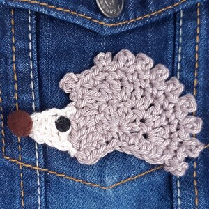 Puede incluir: Un broche de erizo de crochet en tonos marrones y blancos, con una nariz de pompón marrón. El broche está sujeto a una chaqueta vaquera.