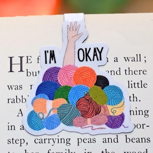 May include: A bookmark with a design of colorful yarn balls, a hand, and the words "I'M OKAY". The bookmark is placed on a book page. The yarn balls are in shades of blue, orange, purple, and green.