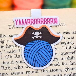 May include: A white bookmark with a pirate hat design, a blue yarn ball, and the text "YAAARRRRRRRRN!". The pirate hat is black with an orange trim and a white skull and crossbones. The bookmark is placed on a book.